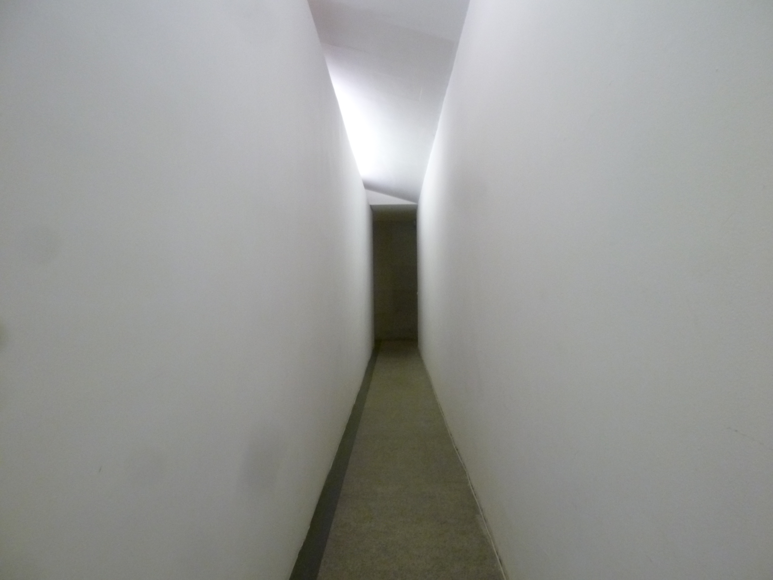 A narrow, overlit service corridor with white walls and a dim passage at the end.